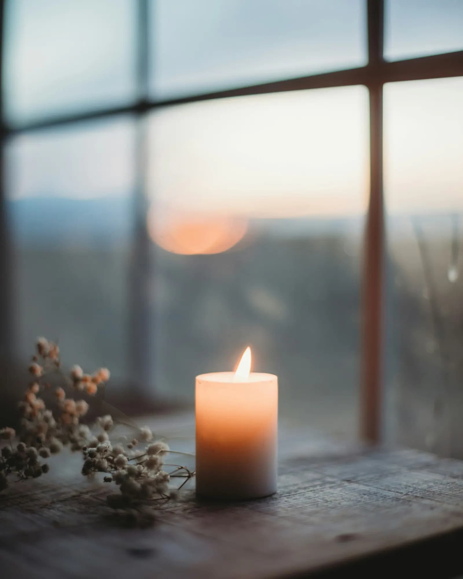 A candle glowing beside dried flowers at a quiet window